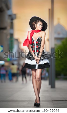 Fashionable lady wearing elegant dress, black hat and red scarf posing outdoor in urban scenery. Full length portrait of young beautiful elegant woman posing in summer city style. Street shot.