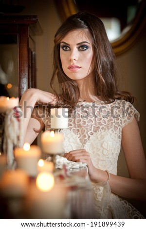 Beautiful sexy woman in white lace dress in vintage scenery with candles. Portrait of long hair brunette girl posing in luxury indoor. Attractive young fashionable female with creative makeup.