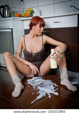 Attractive sexy woman in shirt and socks spilling milk from a bottle. Portrait of sensual girl with long legs sitting on the kitchen floor in comfortable clothes. Indoor shot in modern kitchen