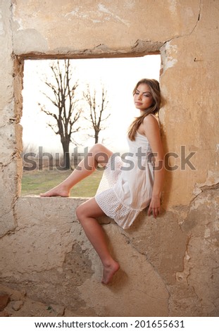 sensual lady in white dress looking to the window and red sun behind her. Lady with long legs standing on the windowsill and looking forward
