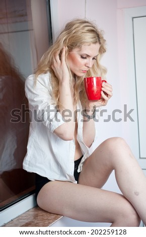 portrait of a young, blond woman, holding a mug with both her hands, wearing a white shirt and black pants, with an expression of being sadness. Woman posing with a big red cup of tea. in  her hands.