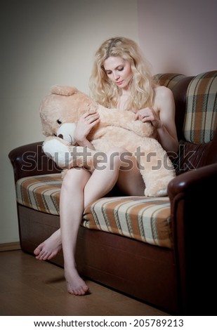 Young blond sensual woman sitting on sofa relaxing with a huge teddy bear. Beautiful girl with comfortable clothes relaxing on the couch with a toy. Attractive blonde in cosy scenery indoor