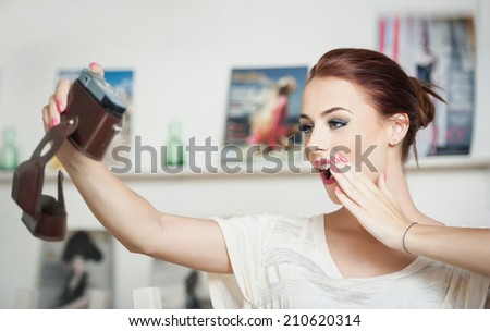 Beautiful, smiling red hair woman taking photos of herself with a camera. Fashionable attractive female taking a self portrait. Selfie, indoor, horizontal. Beautiful redhead taking pictures of herself