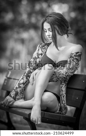Attractive girl in swimsuit sitting relaxed on a bench. Fashionable female model with romantic look posing in park. Beautiful woman in bikini with nice legs sitting daydreaming, black and white.