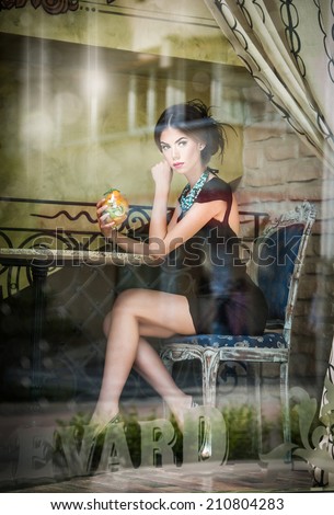 Fashionable attractive young woman in black dress sitting in restaurant, beyond the window. Beautiful brunette posing in elegant vintage scenery with a juice glass. Photo concept through the window