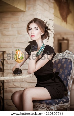 Fashionable attractive young woman in black dress sitting in restaurant. Beautiful brunette posing in elegant vintage scenery with a juice glass. Attractive lady with gloves in luxurious interior