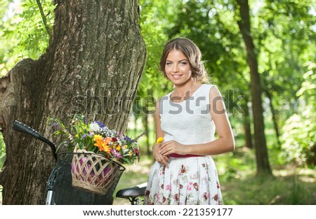 Beautiful girl wearing a nice white dress having fun in park with bicycle carrying a beautiful basket full of flowers. Vintage scenery. Pretty blonde girl with retro look, bike and basket with flowers