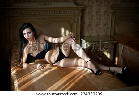 Beautiful sexy brunette young woman wearing black lingerie lying in bed. Fashionable female with attractive body posing provocatively, indoor shot. Sensual girl with high heels shoes on bed in hotel