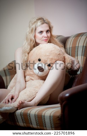 Young blond sensual woman sitting on sofa relaxing with a huge teddy bear. Beautiful girl with comfortable clothes relaxing on the couch with a toy. Attractive blonde in cosy scenery indoor