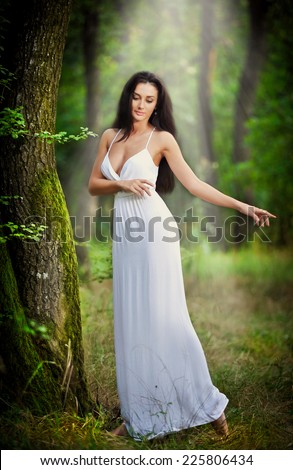 Lovely young lady wearing an elegant long white dress enjoying the beams of celestial light on her face in enchanted woods. Long hair brunette woman looking as a glamorous princess in the forest