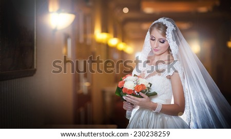 Young beautiful luxurious woman in wedding dress posing in luxurious interior. Bride with long veil holding her wedding bouquet. Seductive blonde bride with fashionable gown, indoor shot