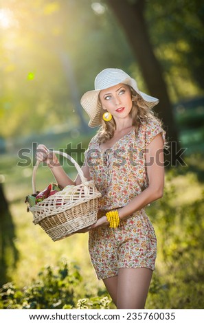 Attractive young woman in a summer fashion shot. Beautiful fashionable young girl with straw basket and hat in park near a tree in sunshine. Blonde woman posing in forest in a sunny day, outdoor shot