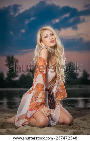 Gorgeous blonde woman in transparent blouse posing provocatively in front of a beautiful sunset. Fair hair girl baring her shoulder and legs in front of lake on cloudy sky background.