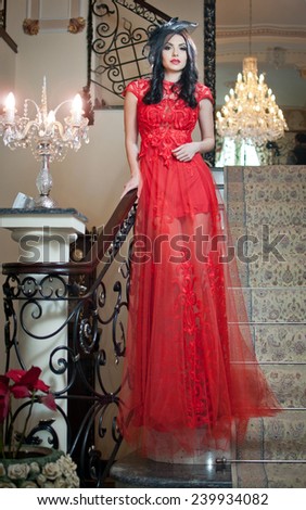 The beautiful girl in a long red dress posing in a vintage scene. Young beautiful woman wearing a red dress in an old hotel. Sensual elegant young woman in red long dress indoor shot.