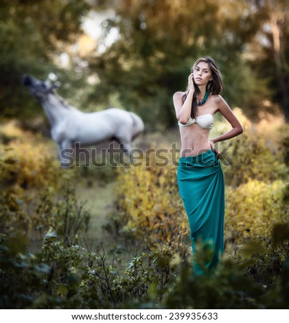 Young women in a blue long skirt and white bra at sunset in forest with a white horse in background .Beautiful young woman with long hair in garden with wild horse. Girl and horse in the field