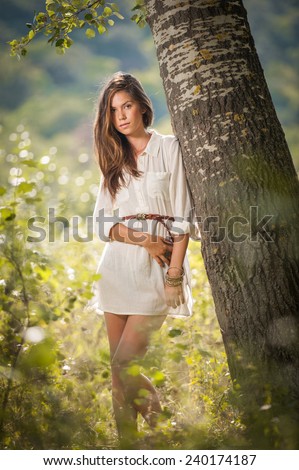 Attractive young woman in white short dress posing near a tree in a sunny summer day. Beautiful girl enjoying the nature in a green forest. Portrait of sensual female in white daydreaming in a meadow