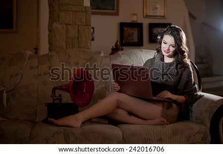 Beautiful young woman sitting on sofa working on laptop having a red gramophone near her, in boudoir scenery. Attractive brunette girl with long hair and long legs laying down on couch with a laptop