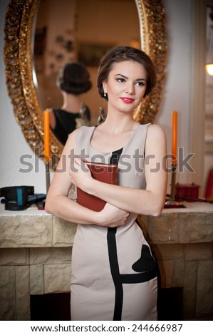 Beautiful sexy woman near a fireplace in vintage scenery. Portrait of girl in slim fit dress holding a book with a large mirror on background. Attractive brunette female  in sensual scenery.