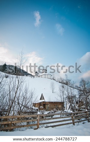View of Romanian small church on hill covered with snow. Winter landscape with orthodox church over blue sky and wooden fence in foreground. Traditional village - Romanian countryside, Moeciu.