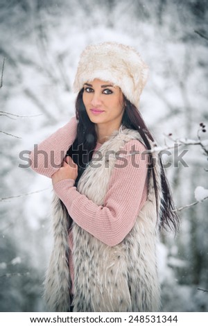 Woman with white fur cap and sheepskin  smiling enjoying the winter scenery in forest. Side view of happy brunette girl posing in winter landscape. Beautiful young female on winter background