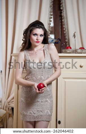Beautiful sexy woman in nude colored lace dress in vintage scenery holding a red apple. Long curly hair brunette young women with makeup in luxury indoor intending to eat a bright red apple