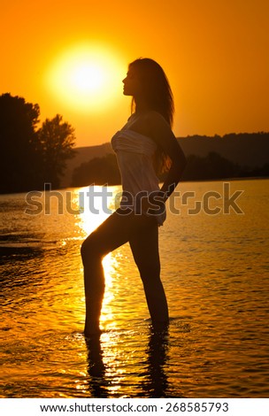 Silhouette of young beautiful woman in the river over sunset sky. Female perfect body contour at beach in twilight scenery, outdoor shot. Sensual girl in water with sun rising behind her.