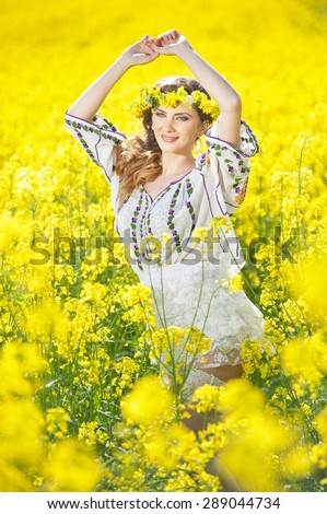Young girl wearing Romanian traditional blouse posing in canola field, outdoor shot. Portrait of beautiful blonde with flowers wreath smiling in rapeseed field