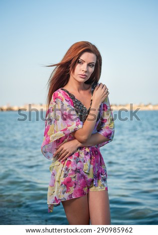 Young sexy red hair girl in multicolored blouse posing on the beach. Sensual attractive woman with long hair, summer shot at sea shore. Perfect body female, holiday concept turquoise water background