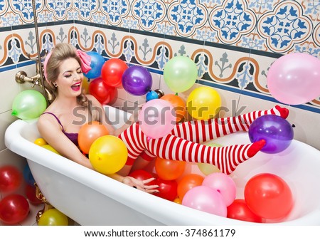 Blonde woman with sunglasses playing in her bath tube with bright colored balloons. Sensual girl with white and red striped stockings having fun in bathroom, covered with balloons