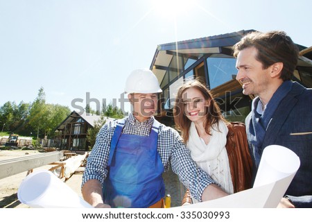 Worker shows construction plan to young couple outside house under construction
