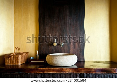 Modern bathroom interior with original white stone sink, clock, wooden baskets and soap dispenser. Wooden interior of spa in yellow and gold colors. Selective focus, only part of bathroom is in focus