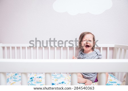 A baby wakes up in her crib crying and screaming under a cloud on the wall.