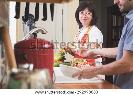 An older woman with short, dark-haired smiles at a younger man who is helping her prepare vegetables in the kitchen.