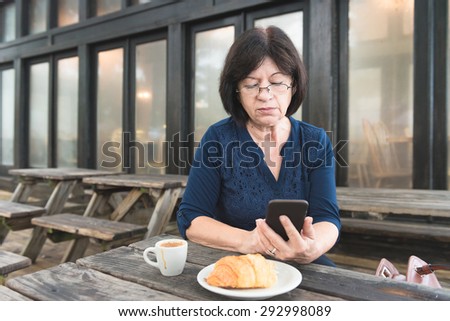 An older woman sits at a wooden table reading on her mobile device while drinking coffee and eating a croissant..