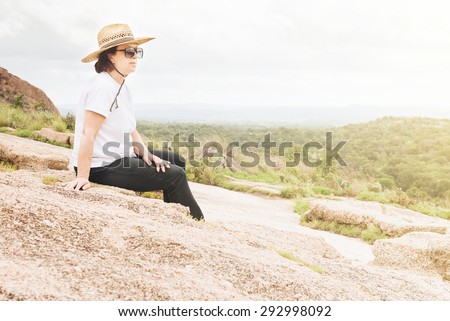 A mature woman with sunglasses and floppy hat sits on a rock outcropping enjoying the green view below.