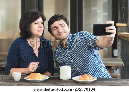 A dark-haired man and an older woman who look like family take a selfie together at a rustic table outdoors before they drink coffee and eat croissants.