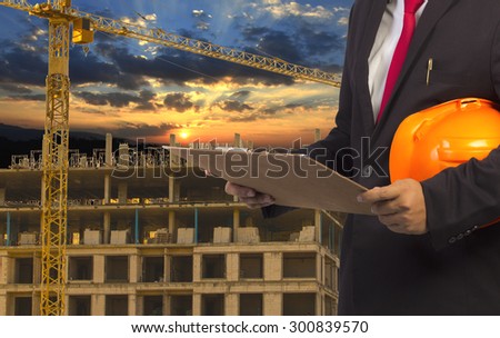 engineer holding orange helmet for workers security on background of new highrise apartment buildings and construction cranes on background of evening sunset cloudy sky Silhouette Crane lifts load