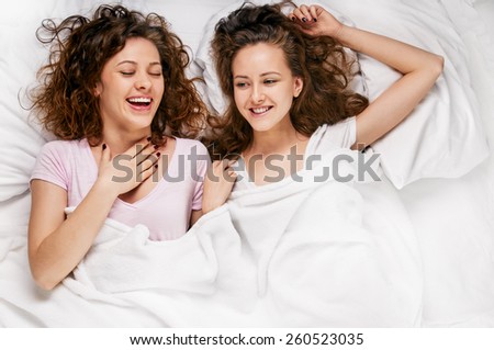 friendship, happiness - two smiling girls whispering gossip, white bed from above