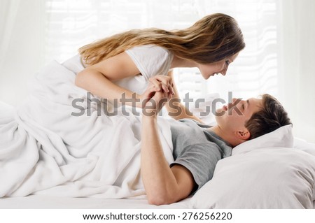 Sexy young couple kissing and playing in bed on light window background