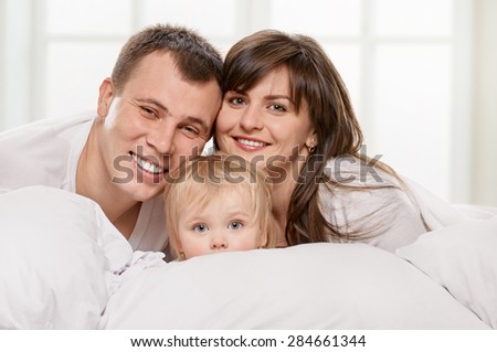 A young family with little daughter on bed in the bedroom on light window background