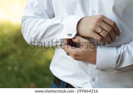 Male puts cufflink. Man dressed in white shirt standing over green nature background. Groom\'s hands in wedding day.