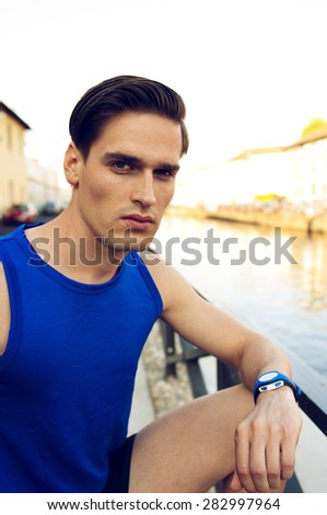 Tired athletic man resting after work by the river. He is wearing blue shirt and sport watch