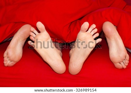 Close up of feet of a happy couple in bed. Two pairs of male and female feet seen from under the red blanket