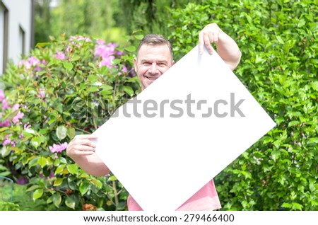 Young cheerful man smiling while holding and showing a blank whiteboard in a green garden with flowering shrubs in a sunny day of spring or summer