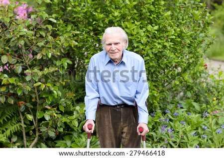 Elderly man with casual clothes using forearm crutches as a mobility aid to walk on the cobblestones of a footpath in a green park or yard, full length
