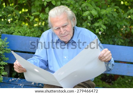 Elderly man sitting alone on a blue wooden bench in the park while holding a blank big white paper sheet and looking at it as at an empty newspaper, in spring or summer, portrait
