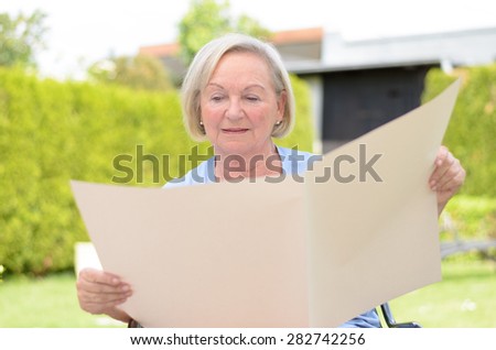 Elderly woman sitting in the park while holding a blank big ocher paper sheet and looking at it as at an empty newspaper, in spring or summer, portrait