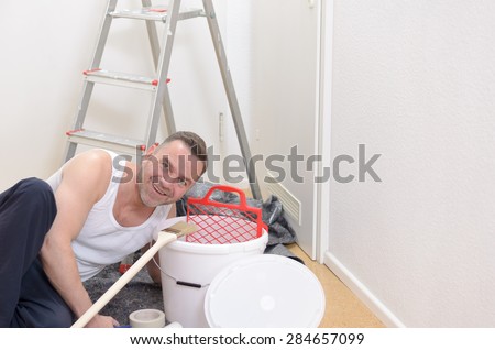 Muscular man doing DIY renovations kneeling down amidst tubs of paint , rollers, paintbrushes and a stepladder looking up at the camera with a smile