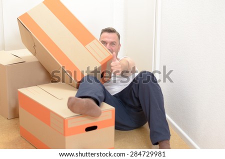 Man moving home relaxing amongst a collection of cardboard boxes packed with his belongings lying on the floor giving a thumbs up of success and approval