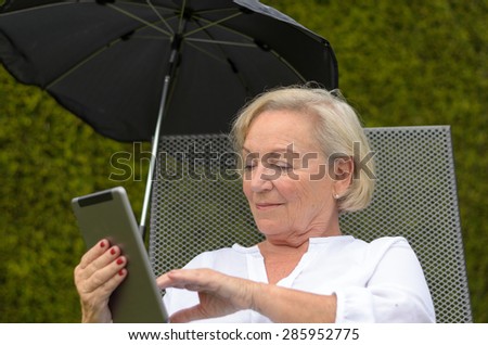 Senior serene woman wearing white shirt and lieing an a camp bed on the green lawn of the garden while using a wireless black tablet PC and showing thumb up, in a warm day of summer
